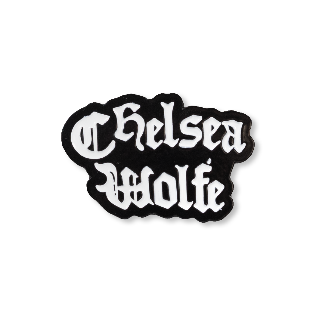 A black and white enamel pin featuring the text 'Chelsea Wolfe' in a white Gothic font on a black background, isolated on a white studio background.